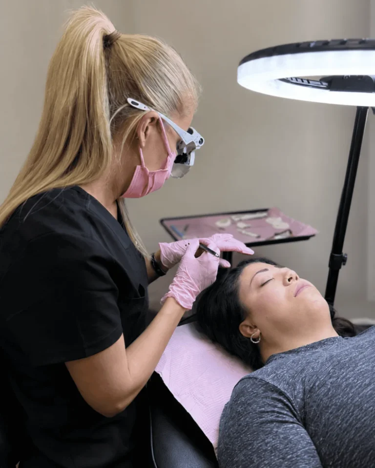 Permanent makeup artist in black scrubs and pink gloves working along a reclining client's brow, with a ring light and instrument tray beside the chair.