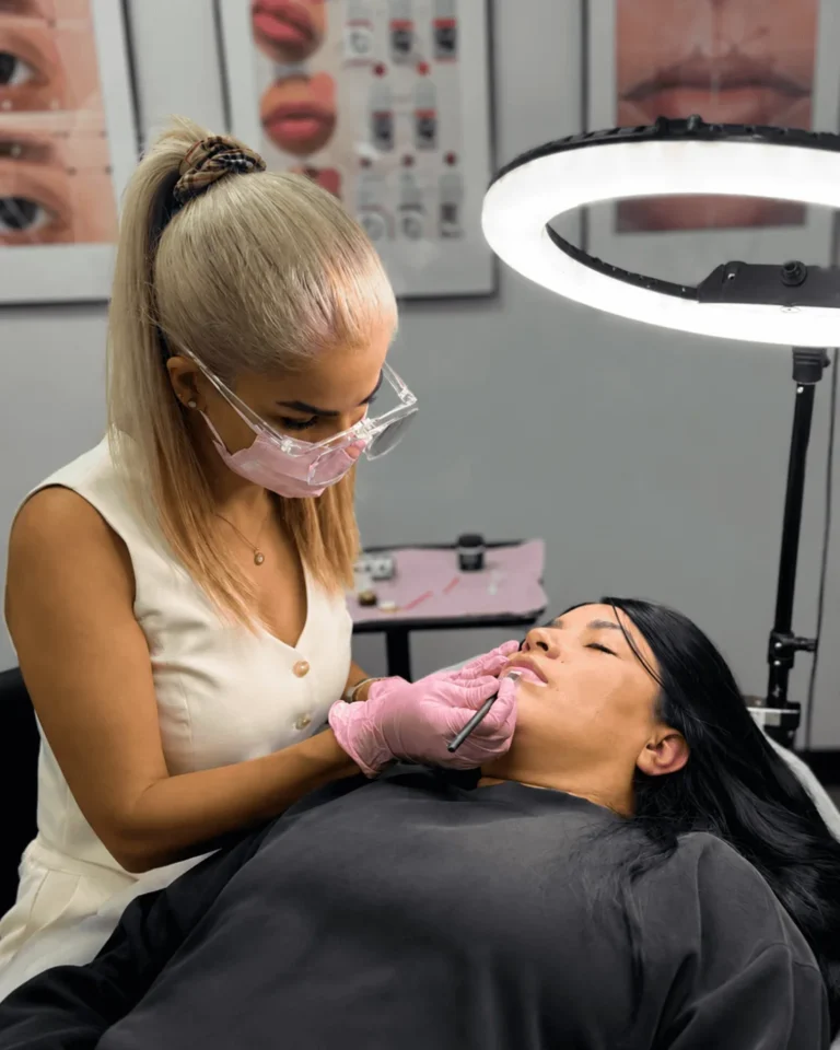 Permanent makeup artist in pink gloves working on a reclining client's lips, with pigment reference charts on the wall behind.
