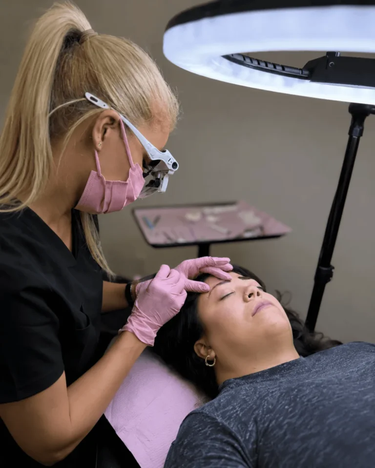 Permanent makeup artist steadying a reclining client's brow with gloved hands under a ring light.