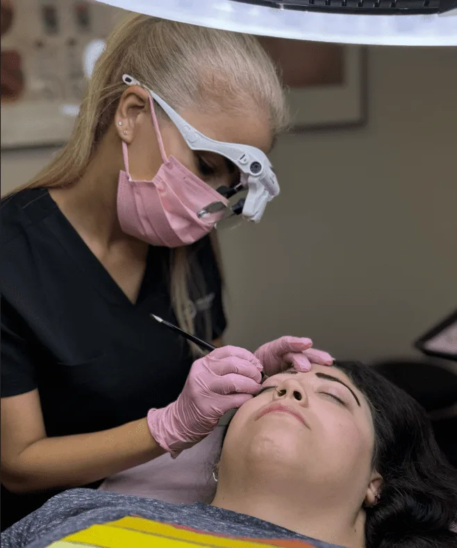 Permanent makeup artist wearing a mask and magnifying loupe, outlining a client's brow while the client rests with eyes closed under a ring light.